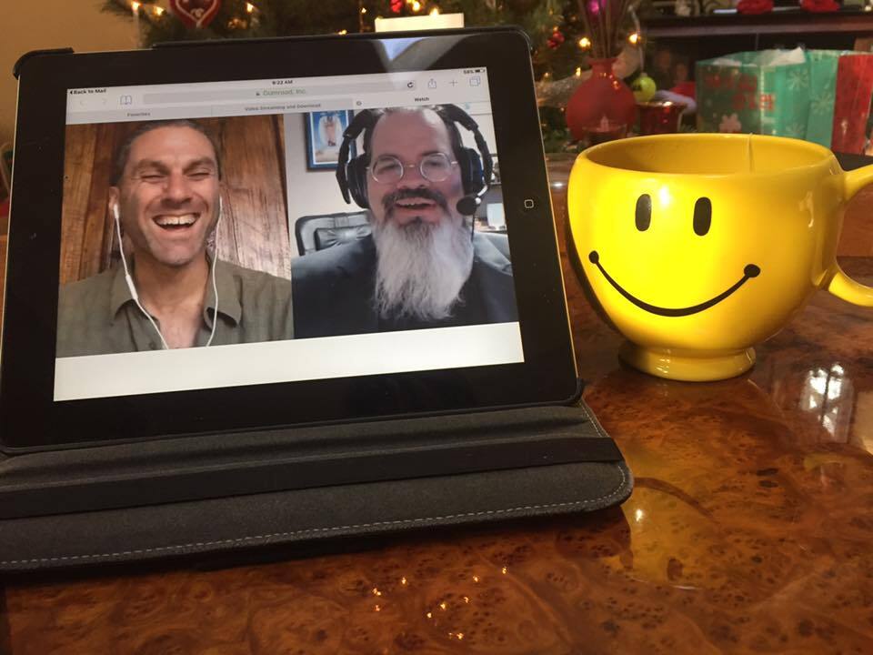 A smiley face coffee cup on a table next to an iPad showing Dave Berman and James Hazlerig laughing during the Laughnosis training course.